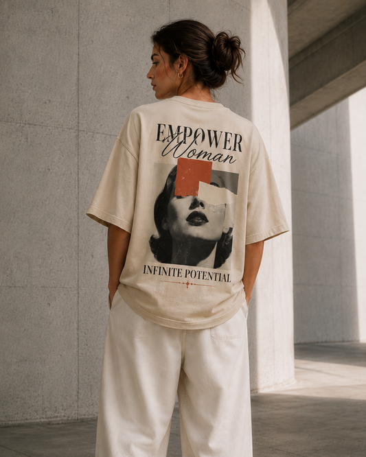 A woman stands with her back to the camera wearing an oversized cream graphic T-shirt and white wide-leg pants in a modern concrete corridor. The back of the shirt features a large graphic print with the words 'EMPOWER Woman' and 'INFINITE POTENTIAL' around a black-and-white portrait with a red geometric accent.