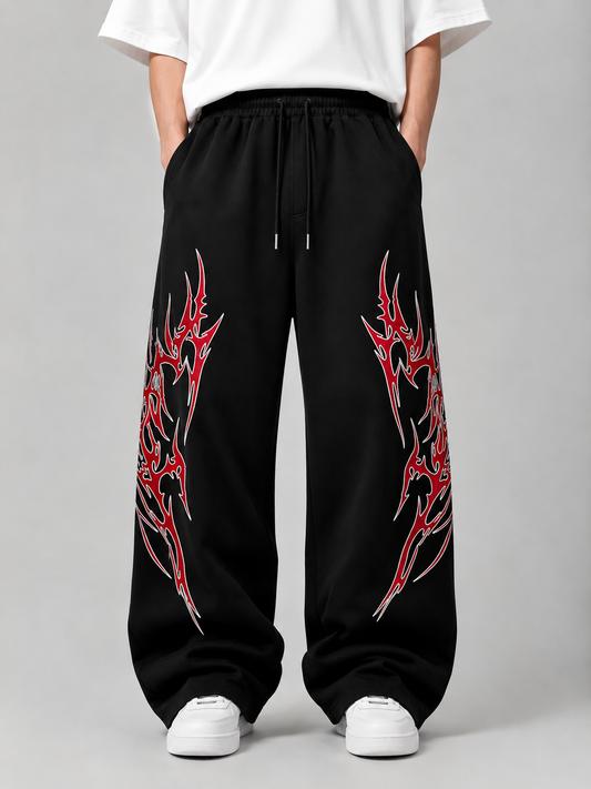 Black wide-leg sweatpants with red and silver tribal-style graphic prints on both legs, shown on a model wearing a white t-shirt and white sneakers against a gray studio background.