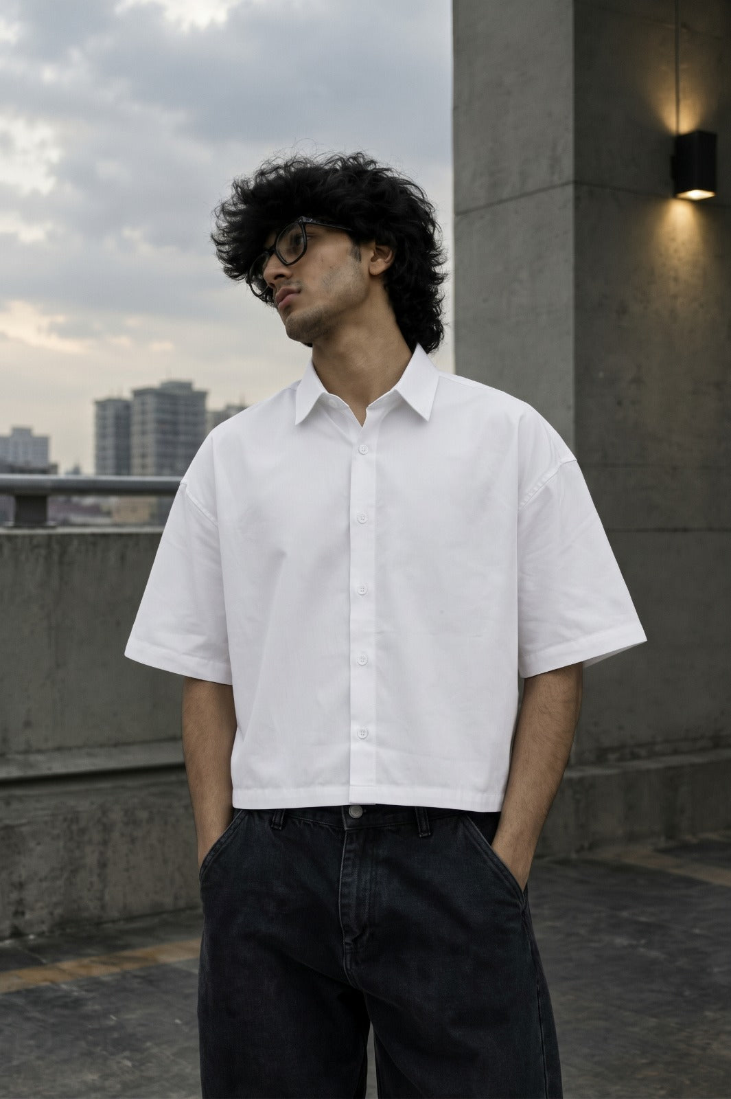 A man wearing a white short-sleeve button-up shirt and dark jeans, standing outdoors on a rooftop terrace with a cloudy sky and city buildings in the background.