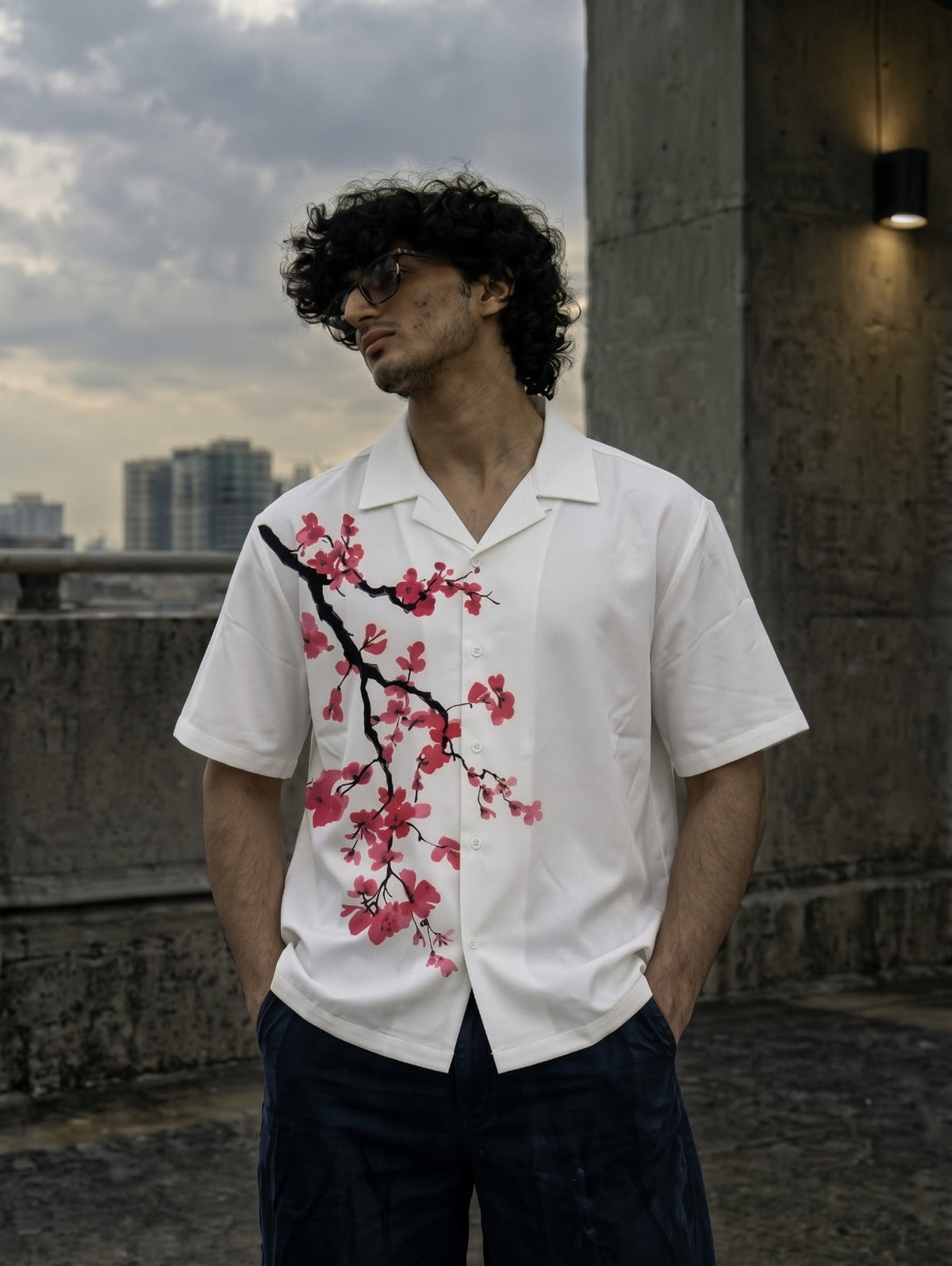 A man wearing a white short-sleeve button-up shirt with a red and black cherry blossom print, standing outdoors against a concrete wall and cloudy sky.