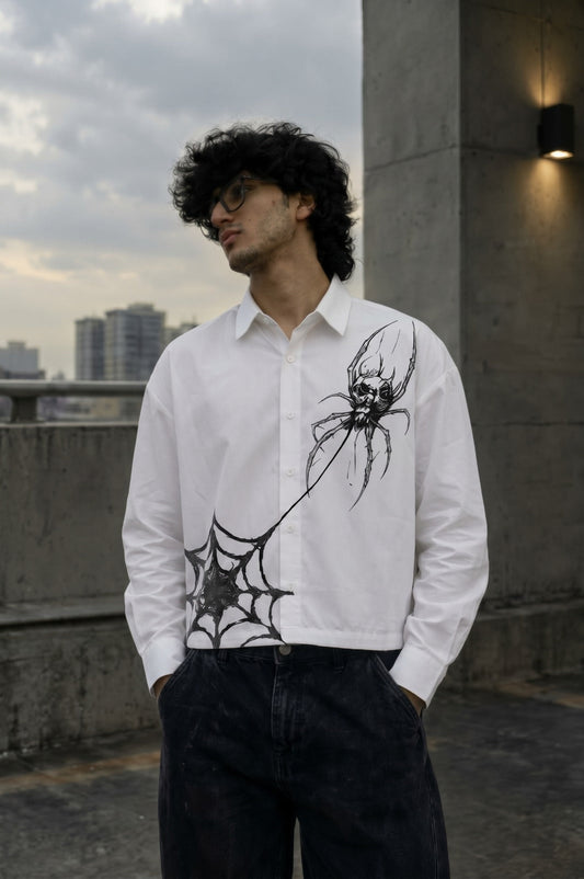 A man wearing a white long-sleeve button-up shirt with a black spider and web graphic print, paired with dark jeans, standing outdoors against a concrete wall and cloudy sky.