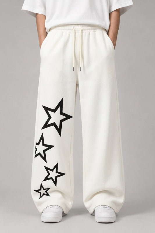 A person wearing loose white drawstring sweatpants with large black outlined star graphics printed down one leg, paired with a white top and white sneakers, against a light gray studio background.