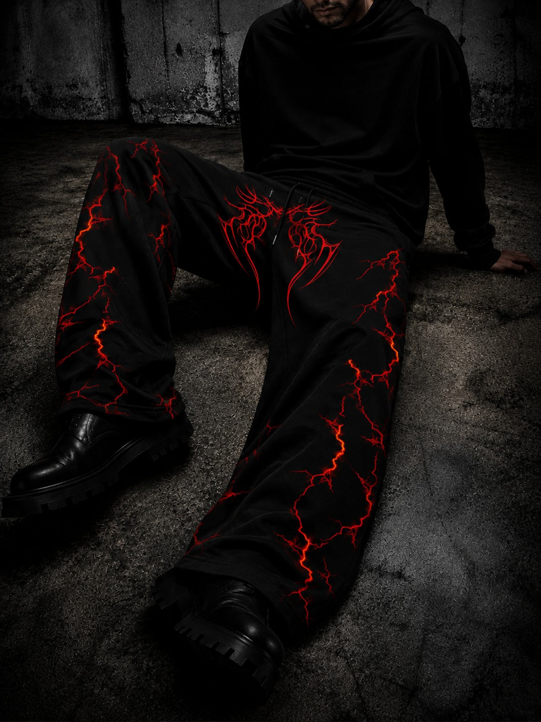 A person wearing black wide-leg pants with a vivid red lightning-bolt graphic pattern, seated on a dark concrete floor.