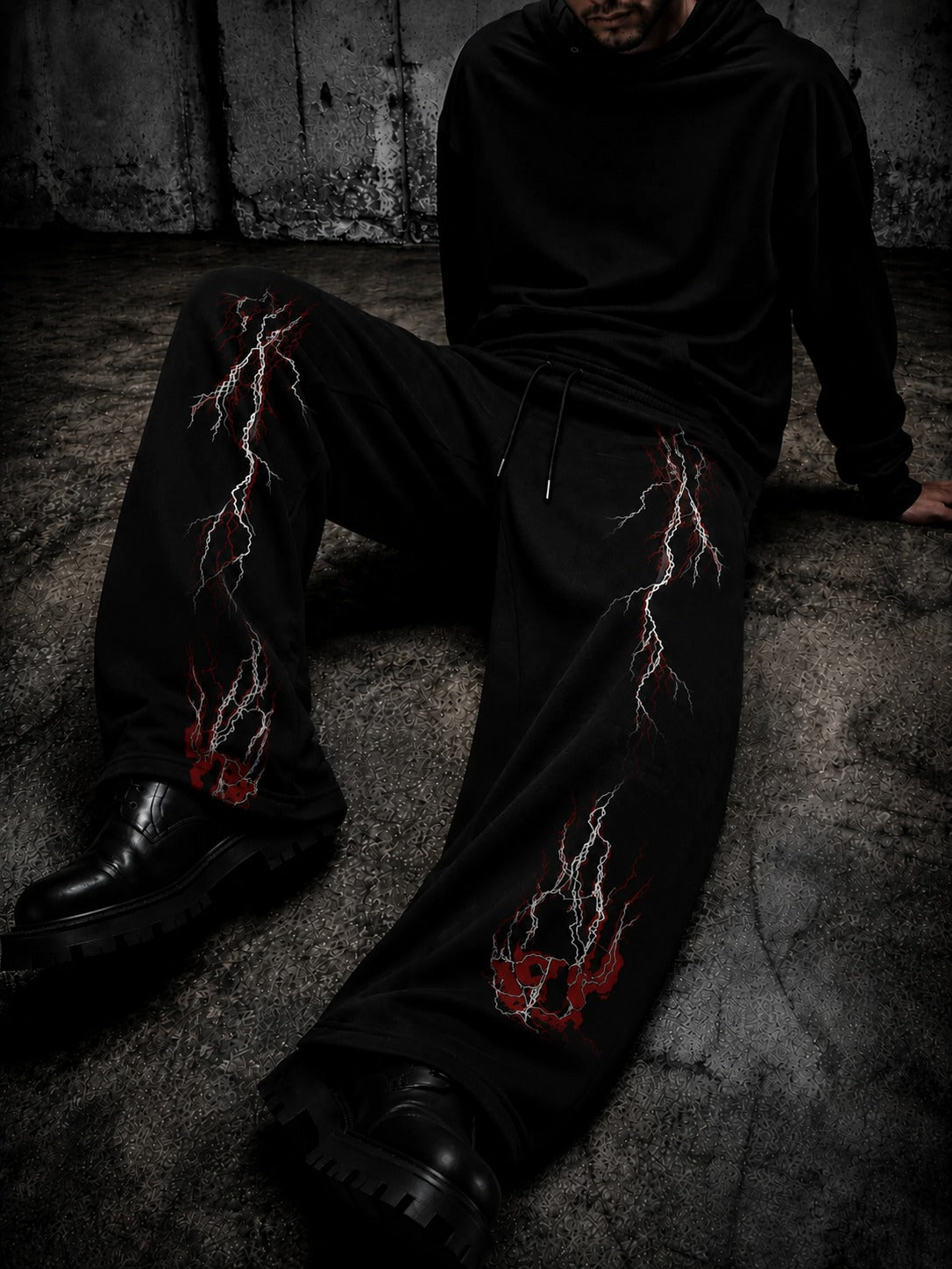 A person wearing black wide-leg sweatpants with red and white lightning-bolt graphics, paired with a black hoodie and black boots, sitting on a concrete floor against a dark textured wall.