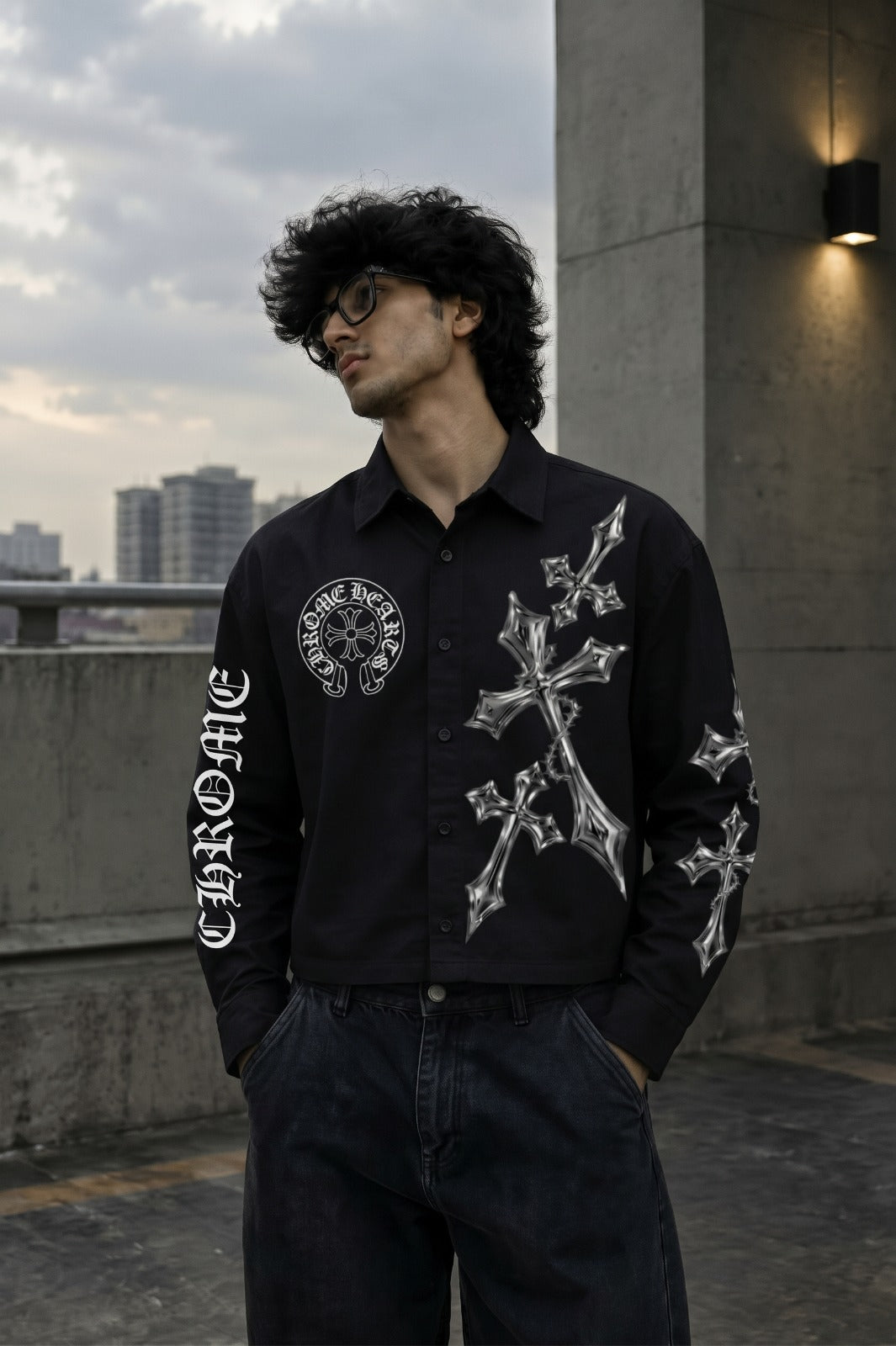 A man wearing a black button-up shirt with white gothic-style cross and circular graphic prints, paired with dark jeans, standing outdoors on a concrete rooftop with a cloudy sky and city buildings in the background.