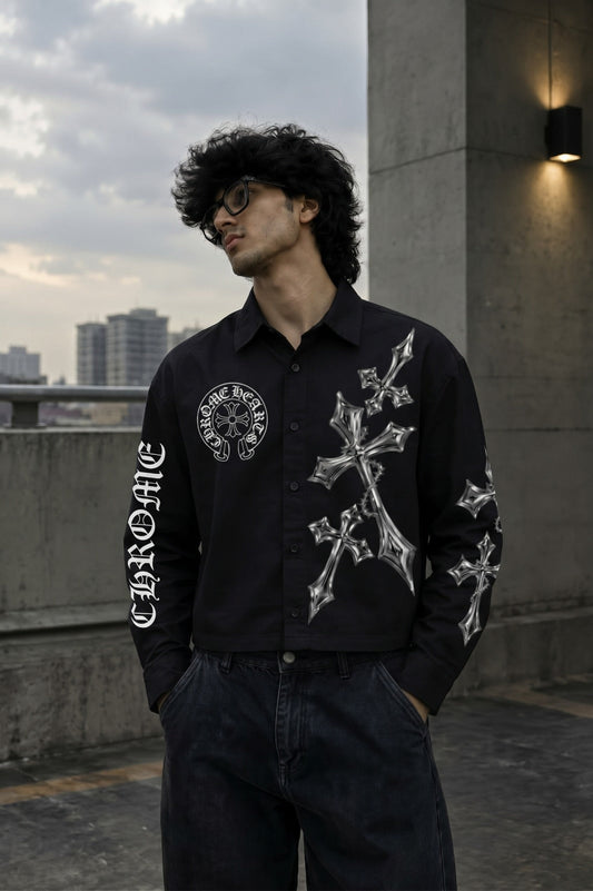 A man wearing a black button-up shirt with white gothic-style cross and circular graphic prints, paired with dark jeans, standing outdoors on a concrete rooftop with a cloudy sky and city buildings in the background.