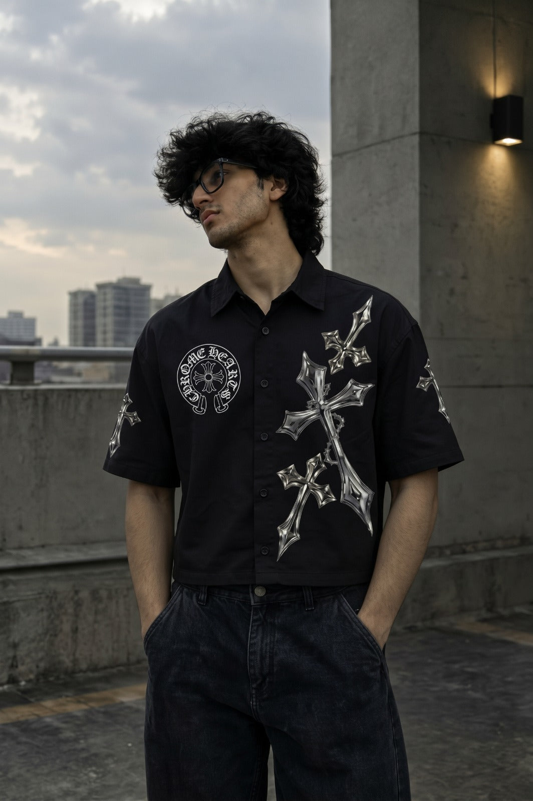 A man wearing a black short-sleeve button-up shirt with large silver cross and circular graphic prints, paired with dark jeans, standing on a rooftop terrace with a city skyline in the background.