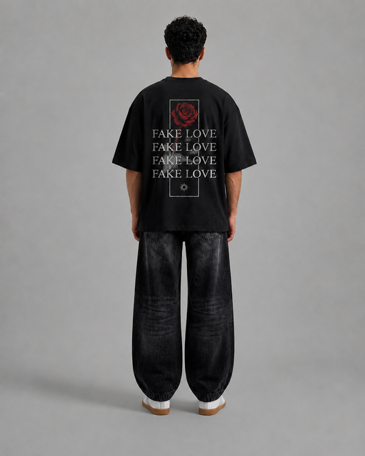A man viewed from behind wearing a black oversized t-shirt with a red rose graphic and the word 'FAKE LOVE' repeated vertically, paired with loose black pants and white sneakers, against a plain gray studio background.