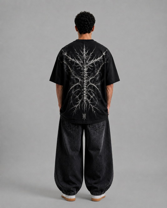 A model viewed from the back wearing a black oversized t-shirt with a detailed white spine and branching graphic print, paired with baggy black jeans, against a solid gray studio background.