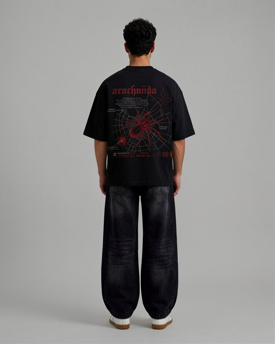 A model viewed from the back wearing a black graphic t-shirt with a red and white spider design and the word 'arachnida' printed on the upper back, paired with dark wide-leg jeans and white sneakers, against a solid gray studio background.