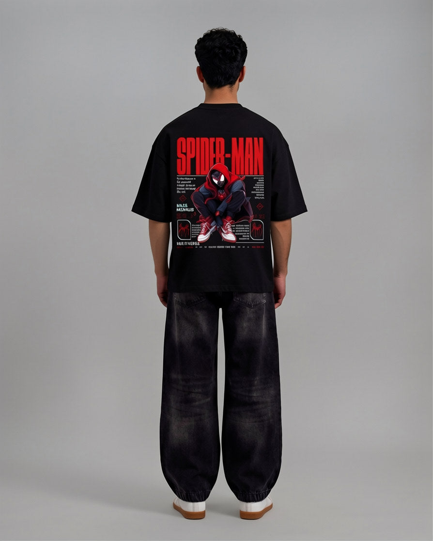 A man viewed from behind wearing a black Spider-Man graphic t-shirt and wide-leg black pants, standing against a neutral gray studio background.