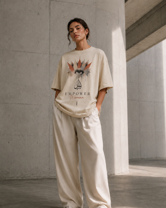 A woman stands in a concrete architectural setting wearing a beige oversized graphic T-shirt with an eye design and the words 'EMPOWER Woman,' paired with matching beige wide-leg pants.
