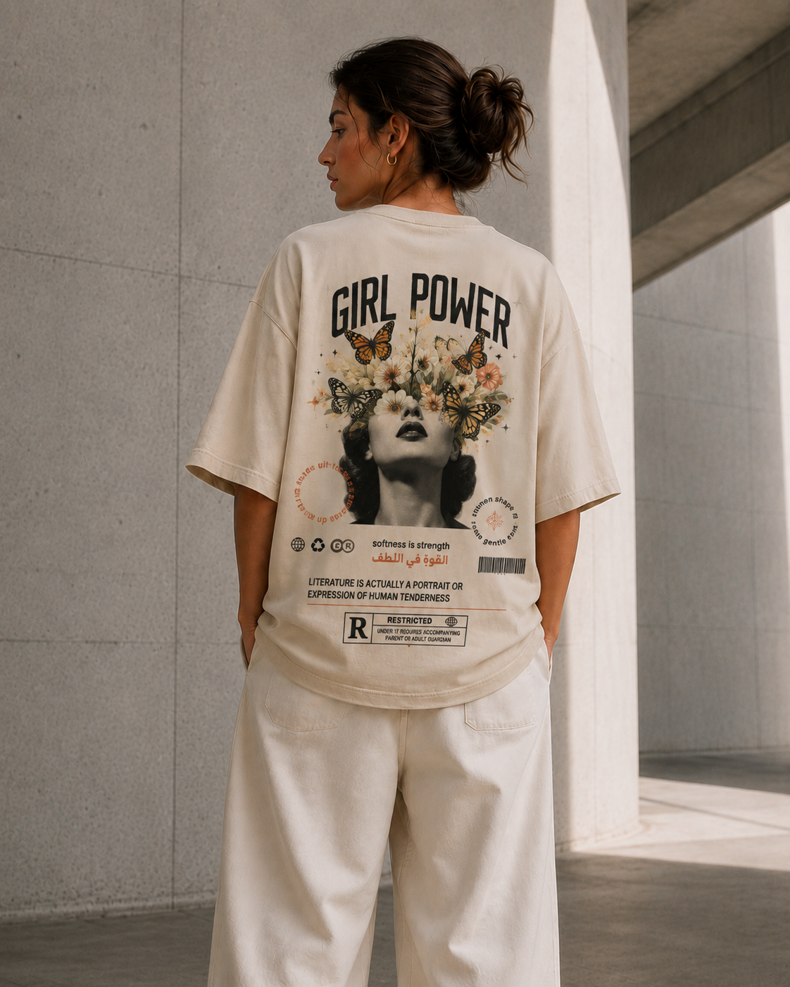 A woman stands in a concrete architectural setting wearing a cream-colored oversized graphic T-shirt and matching cream pants, viewed from behind to show the large back print.