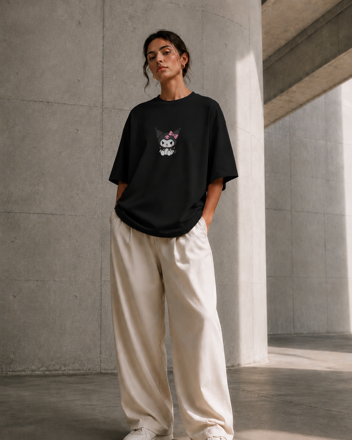 A woman stands in a concrete architectural space wearing an oversized black T-shirt with a small cartoon face graphic on the chest, paired with loose off-white pants and white sneakers.
