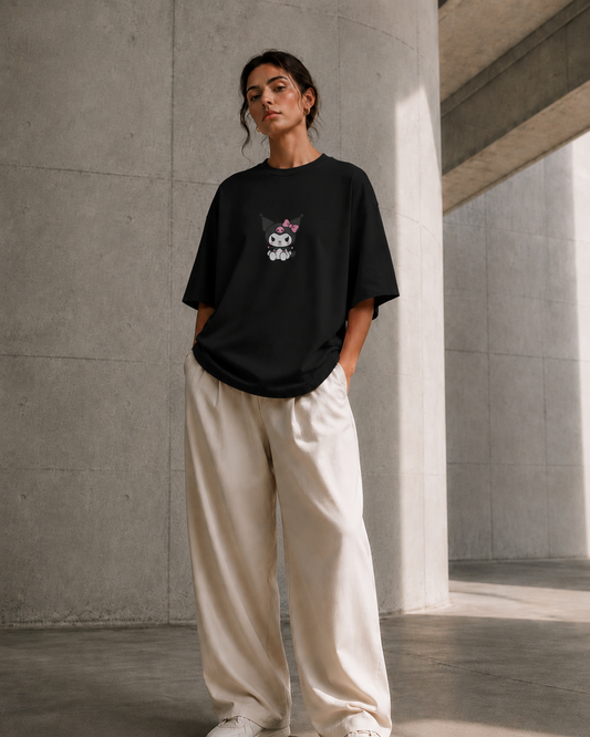 A woman stands in a concrete architectural space wearing an oversized black T-shirt with a small cartoon face graphic on the chest, paired with loose off-white pants and white sneakers.