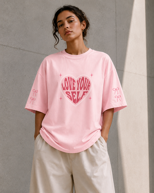 A woman wearing a light pink oversized t-shirt with a pink heart graphic that reads 'LOVE YOUR SELF' and matching bow details on the sleeves, paired with light beige wide-leg pants, standing against a textured gray concrete wall.