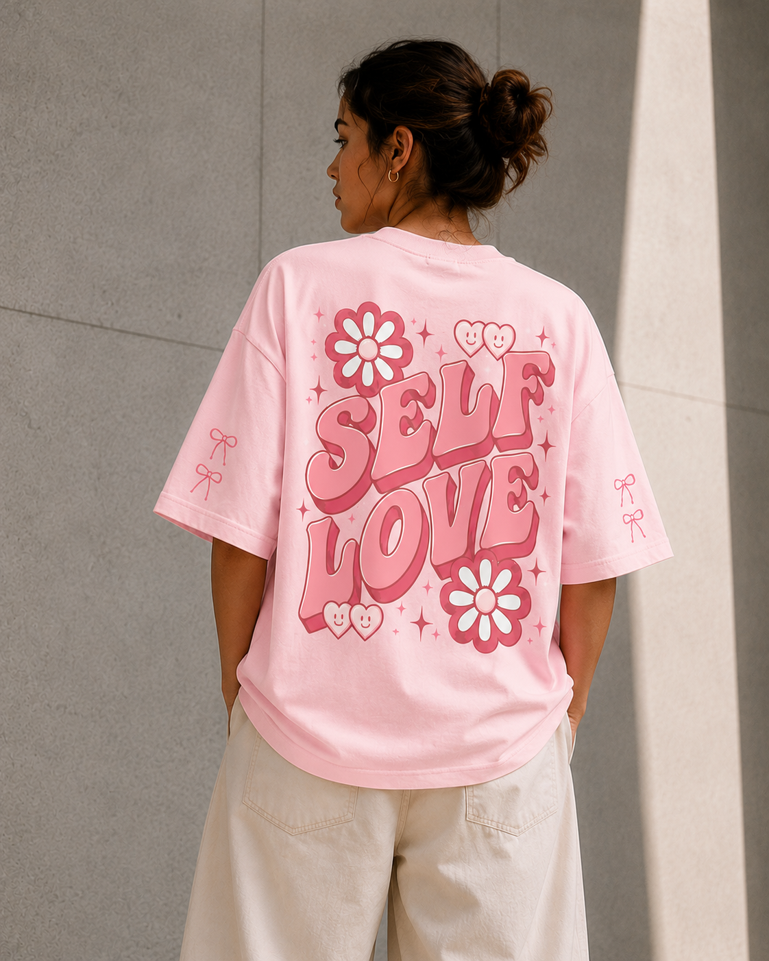 A woman shown from the back wearing a light pink oversized T-shirt with a large pink 'SELF LOVE' graphic, floral motifs, and small bow details on the sleeves, paired with cream pants against a gray concrete wall.