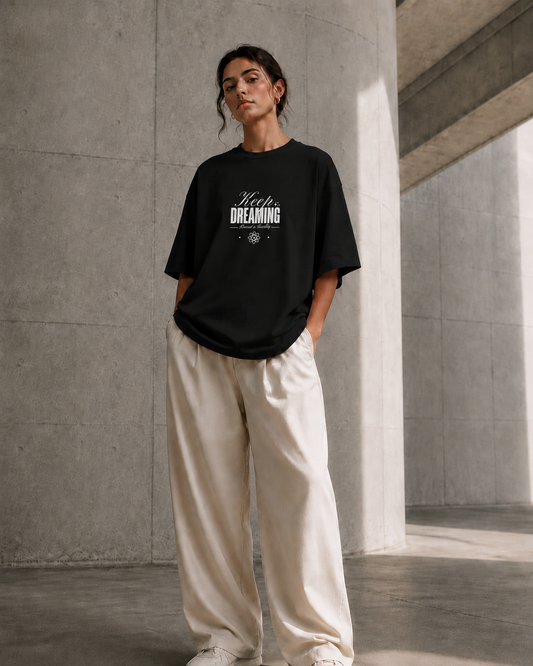 A woman stands in a concrete architectural space wearing an oversized black graphic T-shirt with the words 'Hey DREAMING' printed in white on the chest, paired with loose off-white pants and white sneakers.