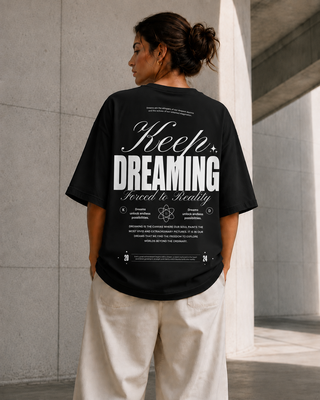 A woman viewed from behind wearing a black oversized T-shirt with a large white graphic print on the back that reads 'Keep Dreaming Forced to Reality,' paired with off-white pants, standing in a concrete architectural setting.