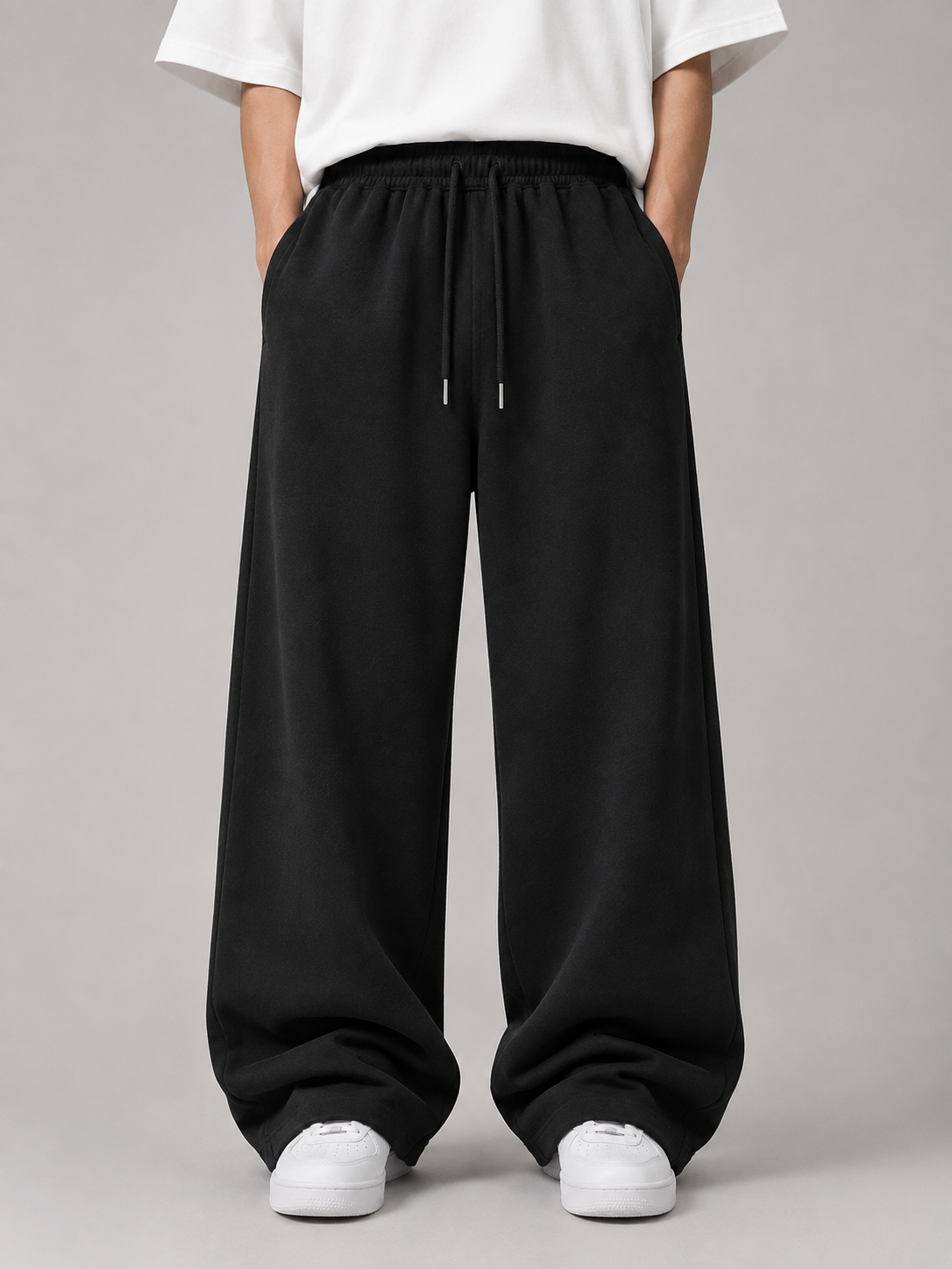 A person wearing loose black drawstring sweatpants with a white t-shirt and white sneakers, shown from the waist down against a plain gray studio background.