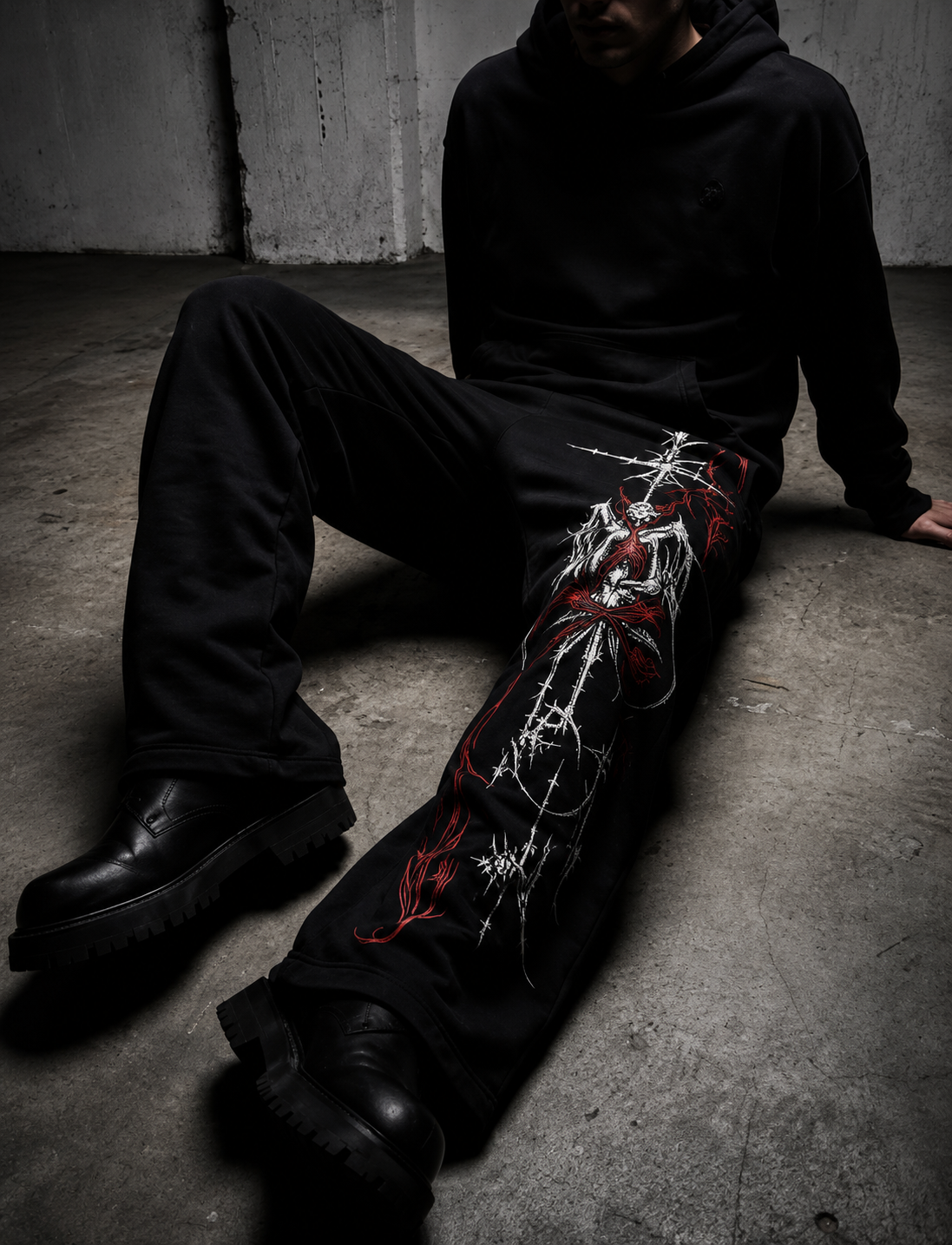 A person sitting on a concrete floor wearing black pants with a large white and red graphic print down the left leg, paired with a black hoodie and black boots.