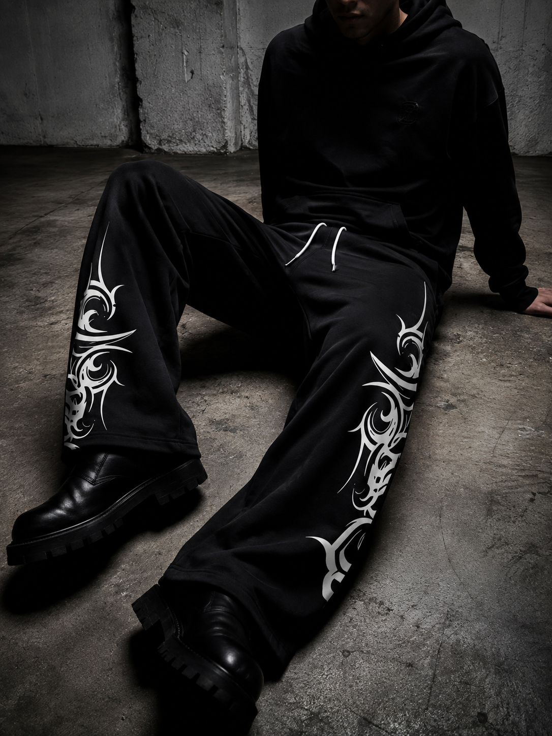 A person seated on a concrete floor wearing black wide-leg sweatpants with large white tribal-style graphic prints down the outer legs, a black hoodie, and black boots in a dark industrial setting.