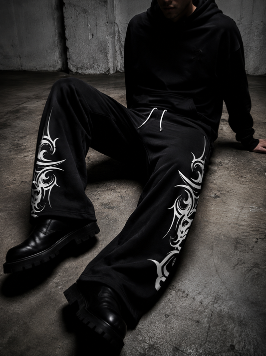 A person seated on a concrete floor wearing black wide-leg sweatpants with large white tribal-style graphic prints down the outer legs, a black hoodie, and black boots in a dark industrial setting.