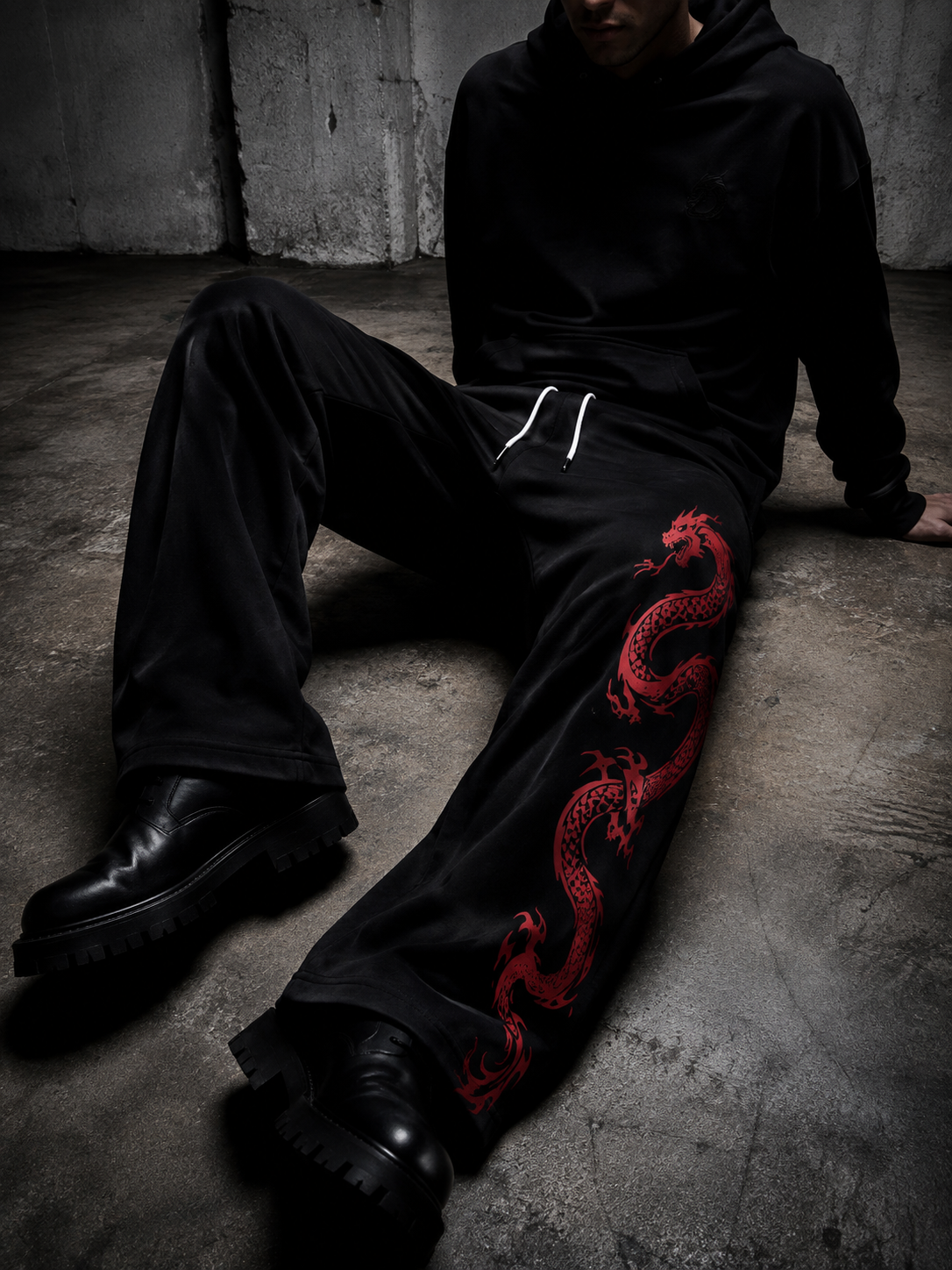 A person wearing a black hoodie and black sweatpants with a red dragon graphic sits on a concrete floor in a dim industrial setting.