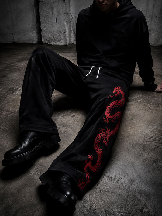 A person wearing a black hoodie and black sweatpants with a red dragon graphic sits on a concrete floor in a dim industrial setting.