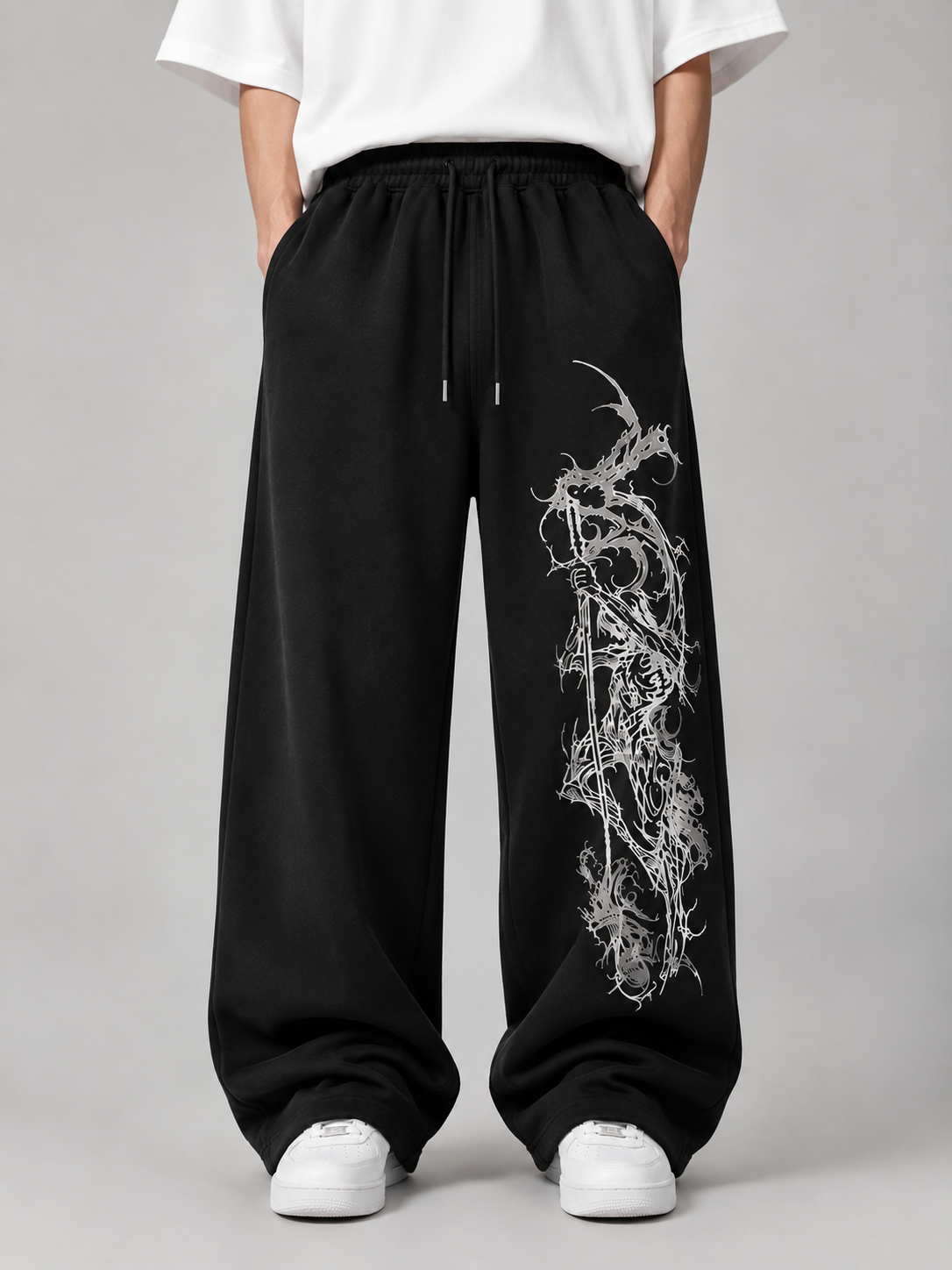 A person wearing black wide-leg sweatpants with a white abstract graphic print down one leg, paired with a white t-shirt and white sneakers, photographed against a neutral gray studio background.