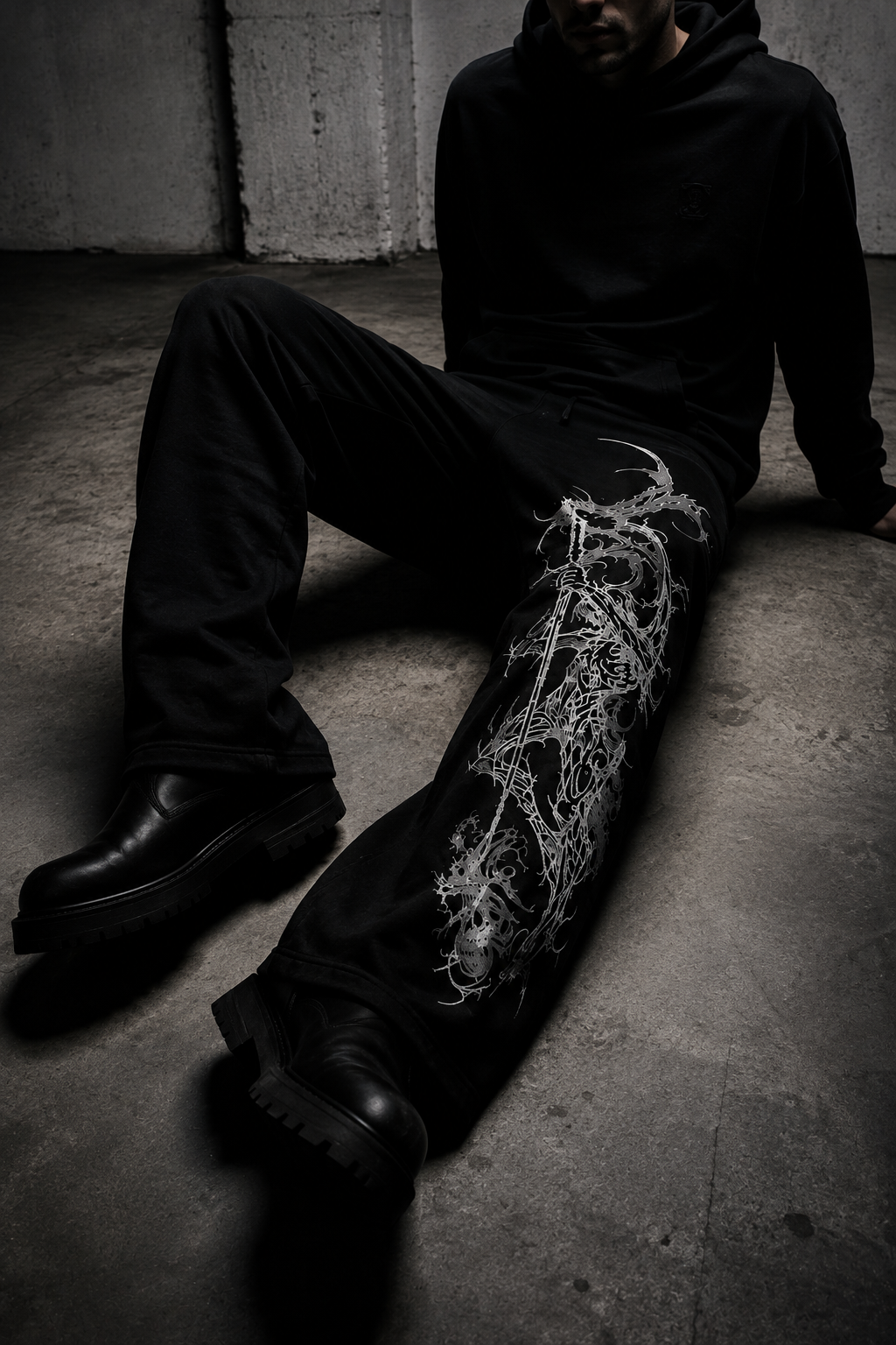 A person sitting on a concrete floor wearing black wide-leg pants with a prominent white barbed wire graphic down one leg, paired with a black hoodie and black boots.