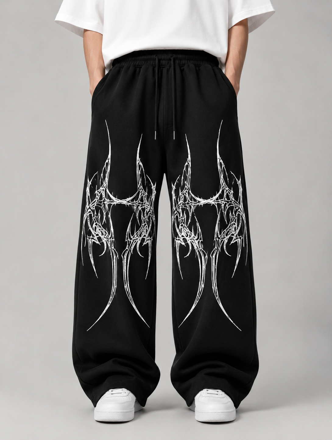 Black sweatpants with a white tribal graphic print on the front, worn by a model in a white t-shirt and white sneakers against a gray studio background.