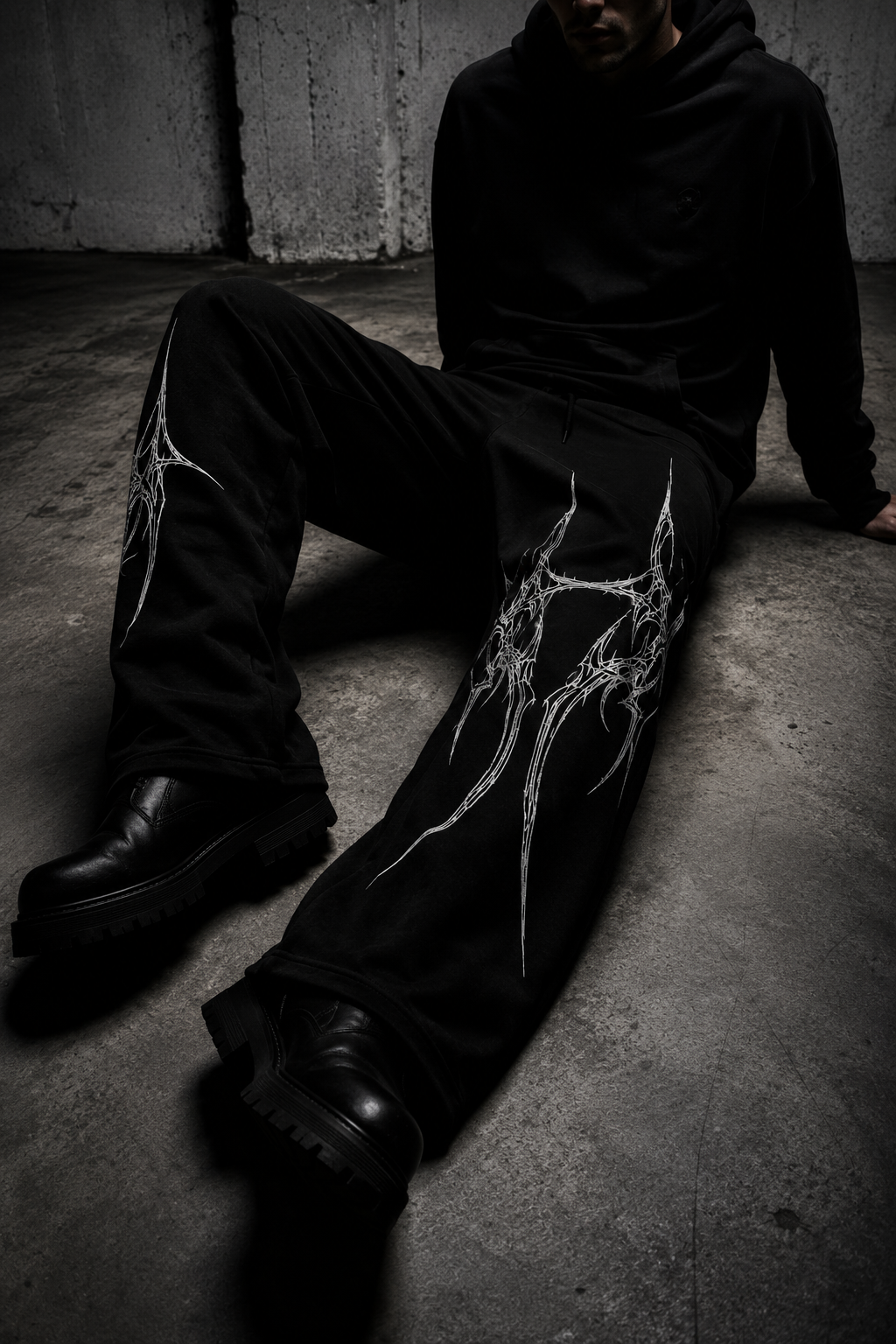A person wearing black wide-leg sweatpants with a large white tribal-style graphic on the left leg, seated on a concrete floor in a dark industrial setting.