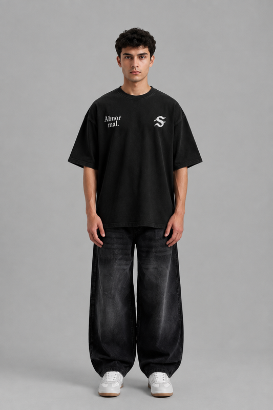 A man stands against a plain gray studio background wearing an oversized black T-shirt with white printed logos on the chest, loose dark jeans, and white sneakers.