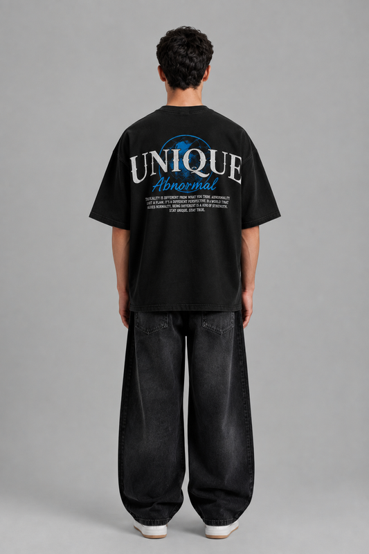 A man viewed from behind wearing a black oversized t-shirt with a large graphic on the back reading 'UNIQUE Abnormal' in white and blue, paired with loose black jeans and white sneakers, against a solid gray studio background.