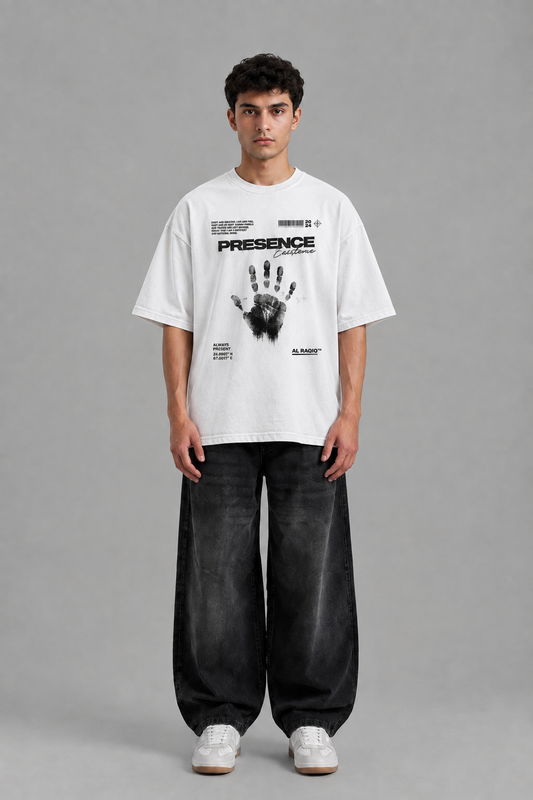 A male model stands against a neutral gray studio background wearing an oversized white graphic T-shirt with black text and a handprint design, loose dark gray baggy jeans, and white sneakers.