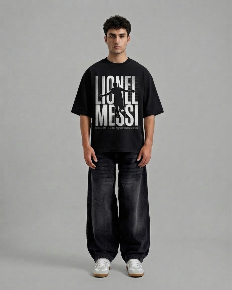 A man stands against a plain gray studio background wearing a black oversized T-shirt with a large white 'LIONEL MESSI' graphic and dark wide-leg jeans with white sneakers.