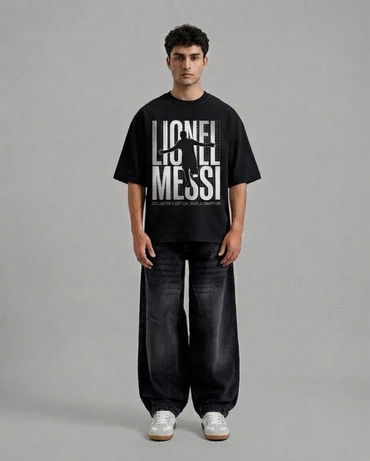 A man stands against a plain gray studio background wearing a black oversized T-shirt with a large white 'LIONEL MESSI' graphic and dark wide-leg jeans with white sneakers.