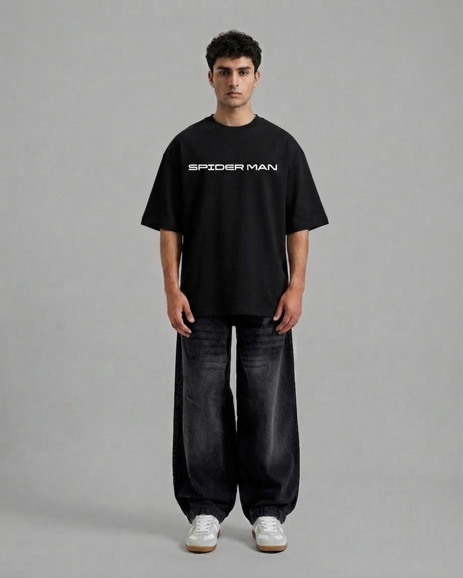 A man stands against a plain gray studio background wearing an oversized black Spider-Man t-shirt with white text, dark wide-leg jeans, and white sneakers.