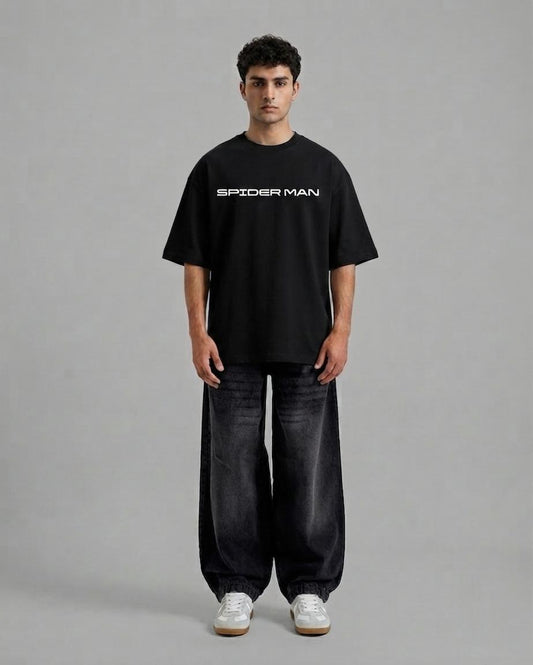 A man stands against a plain gray studio background wearing an oversized black Spider-Man t-shirt with white text, dark wide-leg jeans, and white sneakers.