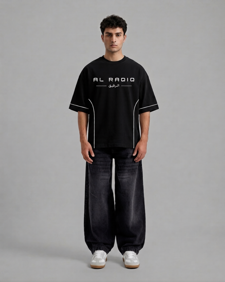 A man stands against a plain gray studio background wearing a black short-sleeve T-shirt with white graphic text reading 'AL RADIO' and white piping details, paired with dark wide-leg jeans and white sneakers.