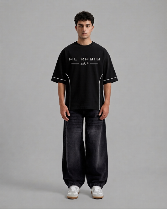 A man stands against a plain gray studio background wearing a black short-sleeve T-shirt with white graphic text reading 'AL RADIO' and white piping details, paired with dark wide-leg jeans and white sneakers.