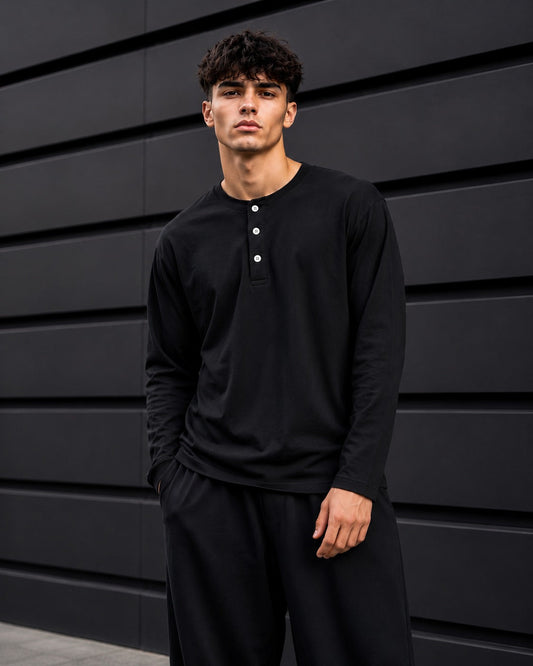 A man wearing a black long-sleeve henley shirt with three white buttons and matching black pants, standing against a dark gray paneled wall.