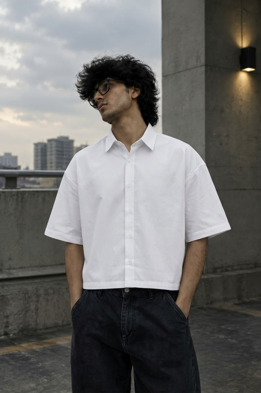 A man wearing a white short-sleeve button-up shirt and dark jeans, standing outdoors on a rooftop terrace with a cloudy sky and city buildings in the background.