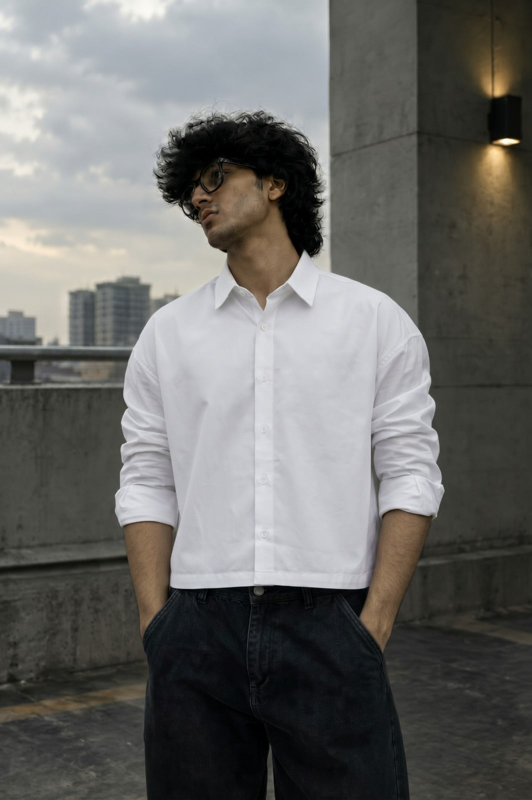 A man wearing a white long-sleeve button-down shirt and dark jeans, standing outdoors on a rooftop with a cloudy sky and city buildings in the background.