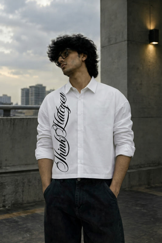 A man wearing a white long-sleeve button-up shirt with a large black script graphic printed vertically down the left side, paired with dark pants, standing outdoors on a rooftop with a cloudy sky and city buildings in the background.