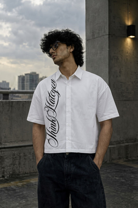 A man wearing a white short-sleeve button-up shirt with a large black gothic script graphic down the left side, paired with dark jeans, standing on an outdoor rooftop with a city skyline in the background.