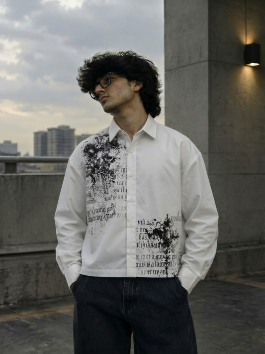 A man wearing a white long-sleeve button-up shirt with a black graphic print on the front, paired with dark jeans, standing outdoors on a rooftop with a cloudy sky and city buildings in the background.