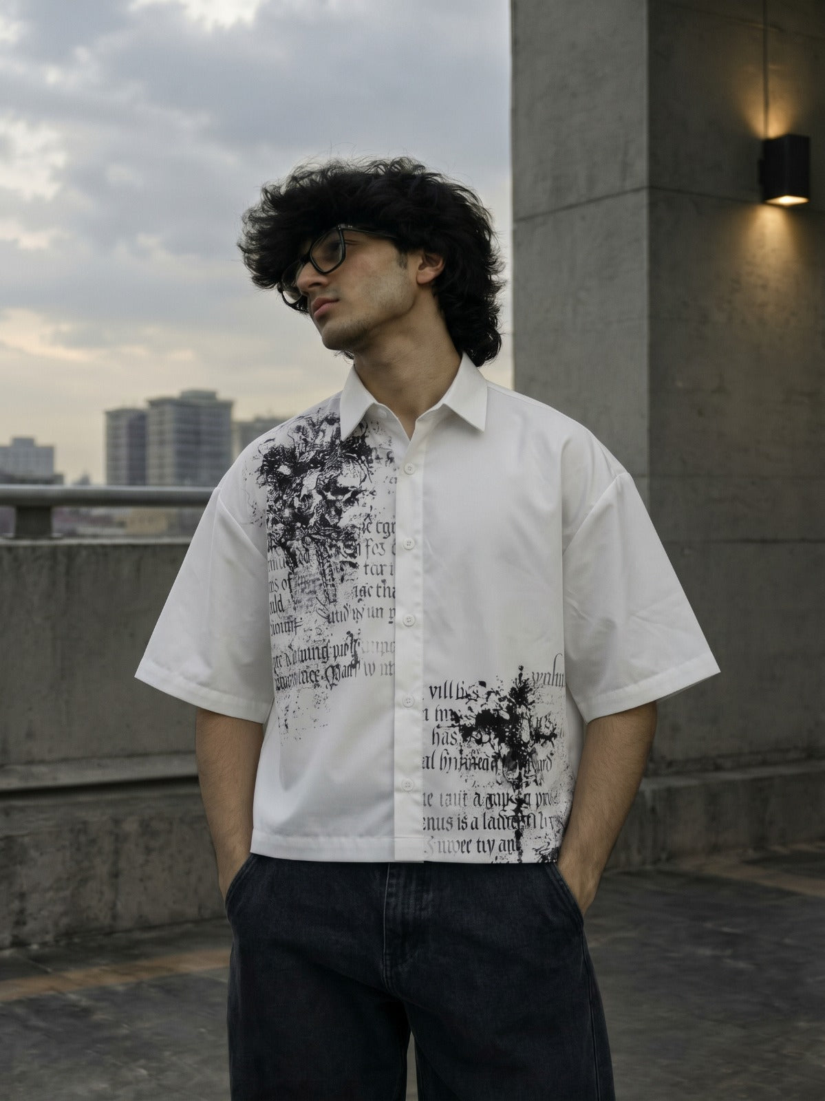 A man wearing a white short-sleeve button-up shirt with black graphic text and paint-splatter style prints, paired with dark jeans, standing outdoors on a rooftop with a cloudy sky and city buildings in the background.