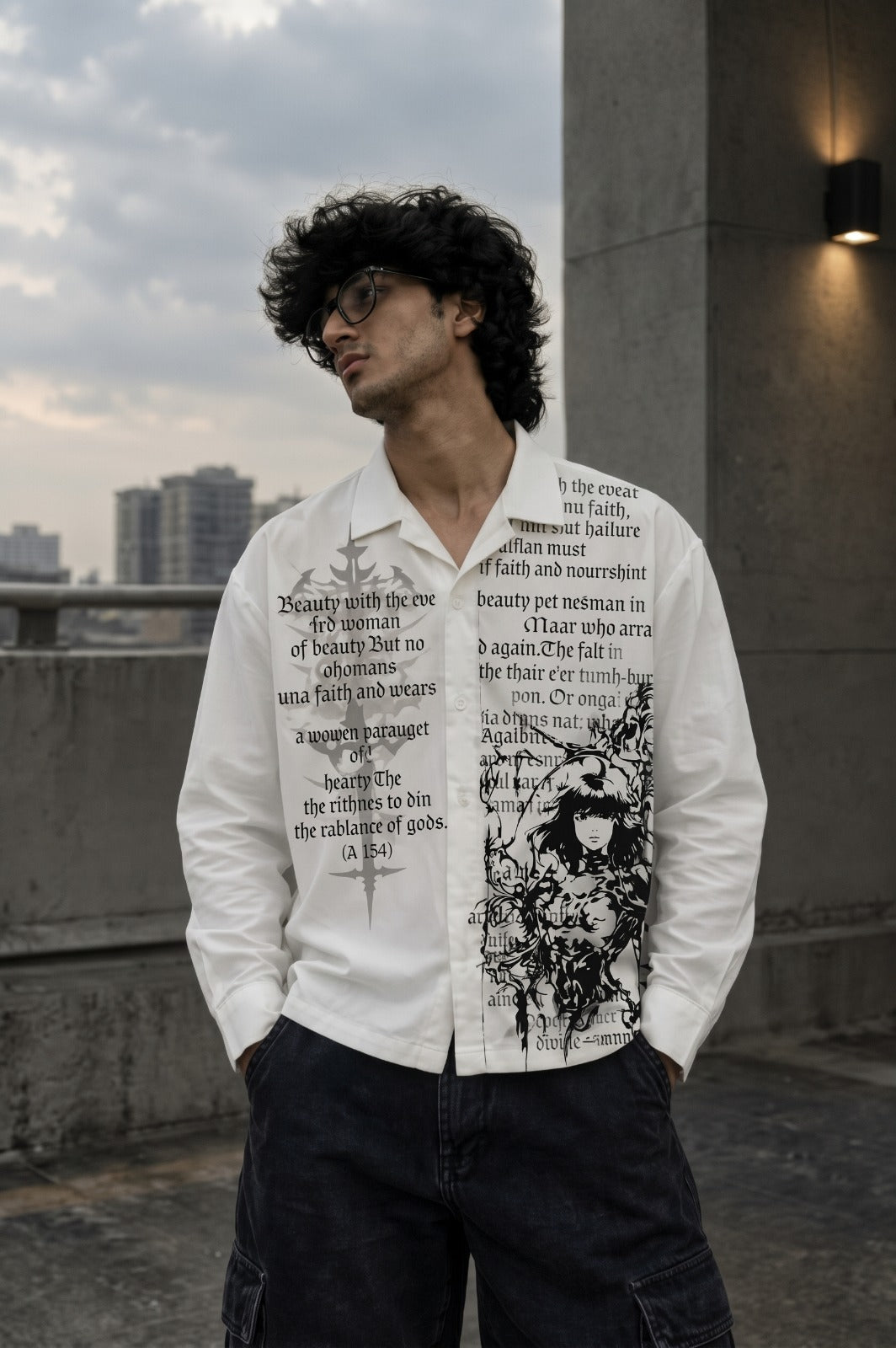 A man wearing a white long-sleeve button-up shirt with black graphic text and illustration prints, paired with dark cargo pants, standing outdoors on a rooftop with a city skyline in the background.
